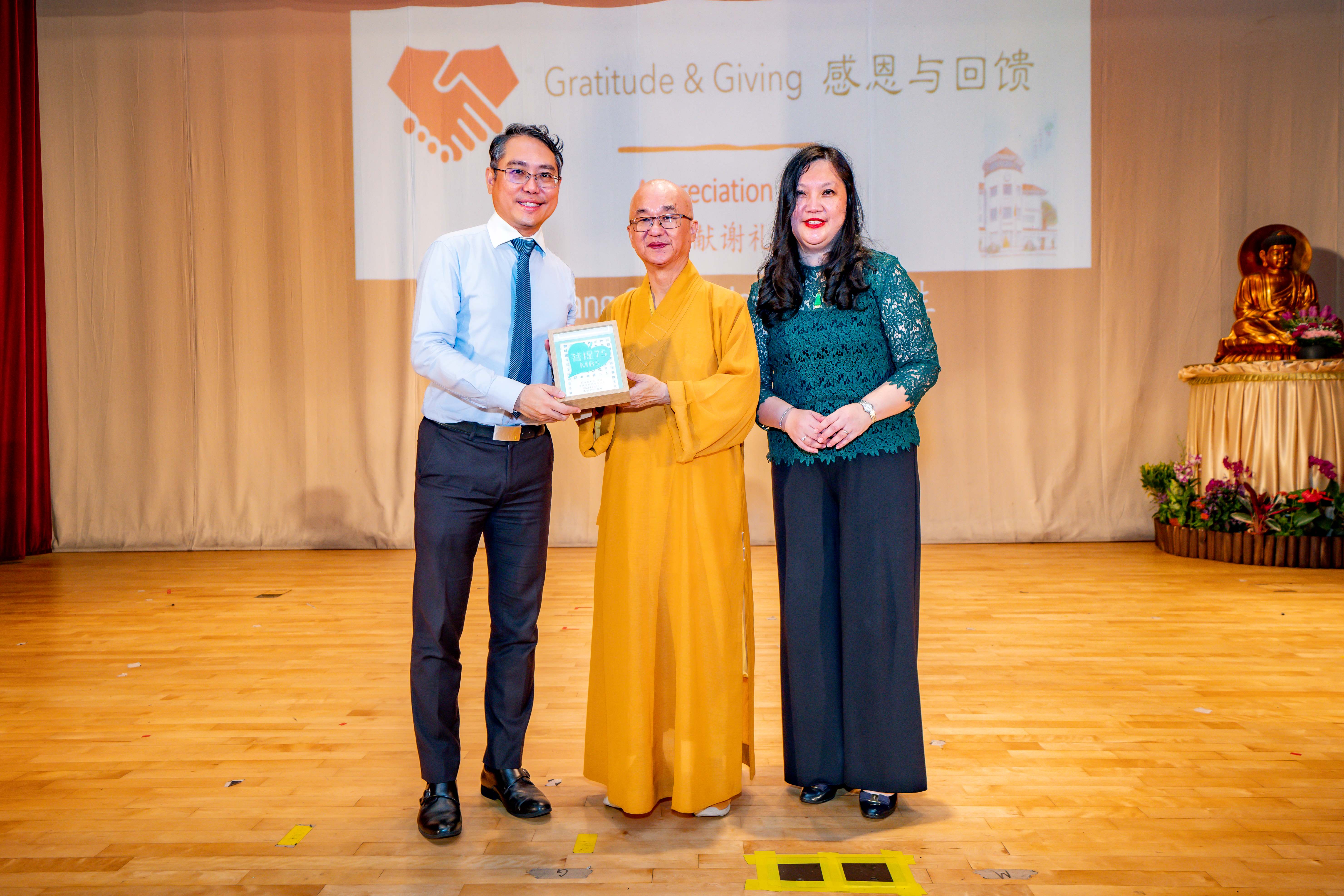 Three people stand on a stage at a Gratitude and Giving event, with a Buddhist monk in yellow robes holding a small gift.